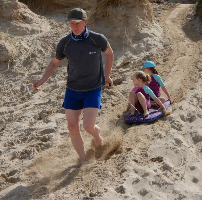 Man towing his two grand children, who are sitting on a body board, down a large sand dune 